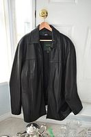 Front view of the Danier men's black leather jacket hanging on a door with a silver doorknob, showing full jacket and collar.