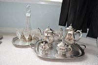 Full view of silverplated tea set on rectangular tray with separate round tray holding glass decanter and glasses