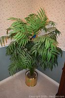 Tall faux plant with green leaves in a brass-colored metal pot against floral wallpaper and blue painted wall.