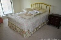 Full view of the brass colored bed frame with floral bedding, bed skirt, and pillows on a carpeted floor near a window.