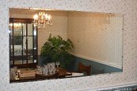 Photo of large rectangular unframed mirror mounted on wall reflecting dining room with chandelier and furniture.