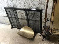 Full view of black mesh fireplace screen with two hinged doors, metal log bin with wear, cast iron tool rack and wooden handled fireplace tools arranged on floor.