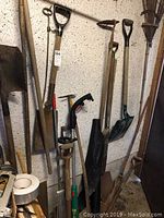 Photo showing assortment of garden shovels, rakes, hand tools, and part of a wooden hand truck.