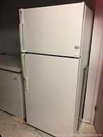 White GE top freezer refrigerator with two doors and vertical handles, standing next to a chest freezer.