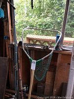 Fishing net leaning against window with can of paint nearby, showing metal frame and green netting on wooden handle.
