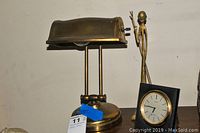 Brass desk lamp, brass warrior statue, and black framed clock grouped together against a white wall.
