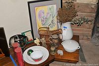 Full lot from a distance showing framed artwork, porcelain jug with flowers, German stein, candles, and other decorative items on wooden table.
