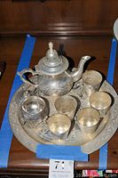 Overview image of Middle Eastern style tin tone tea set on wooden cabinet, showing tea pot, sugar bowl, six tea cups, and round tray with detailed engravings.