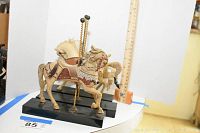 Two carousel horse figurines mounted on black bases with gold-colored twisted poles, showing detailed painted and carved designs.