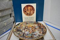 Photo of one decorative plate displayed flat inside a blue presentation box with printed descriptive insert titled 'Jacob's Wedding'. Plate depicts a detailed artistic scene with multiple figures and musical instruments in warm earth tones.