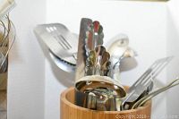 Image shows a close-up view of assorted stainless steel kitchen utensils inside a cylindrical bamboo holder, including tongs, spatula, and forks.