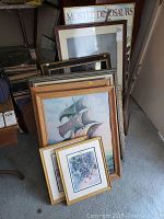 Framed artworks stacked upright, various sizes and styles, wood and gold frames visible