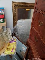 Photo showing large ornate gold framed mirror leaning against wall near wooden furniture with bubble wrap on smaller mirror