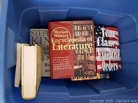 Collection of books placed in a blue storage container, showing titles Merriam Webster's Encyclopedia of Literature, Tom Clancy Executive Orders, Stephen King book.