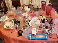 Wide shot of assorted glassware including bowls, plates, cups, pitchers, vases, and decorative ceramics on table covered with orange cloth.