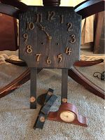 Black wooden WM Gilbert wall clock face with gold numbers and hands, pendulum parts and Seth Thomas mantle clock visible below, showing the worn condition and assembly.