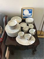 Full lot of English Garden china on an end table showing plates stacked in groups, cups on a tray, and boxed vegetable bowls visible in background.