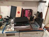 Kitchen countertop with multiple kitchen items including flatware sets, pots, frying pan, knife sets, coffee pod shelf, and storage containers.
