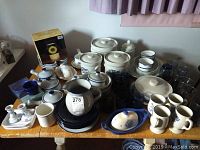 Overview photo showing a large assortment of ceramic kitchenware items primarily Pfaltzgraff brand with blue floral designs, including covered casseroles, bowls, mugs, a pitcher, butter dishes, and glassware on a wooden table.