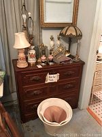 Array of 9 table lamps, including ceramic and glass, placed on and around a wooden cabinet along with 2 hanging brass-looking fixtures and a lampshade.