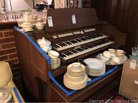 Photo of china dishware including multiple stacks of plates, bowls, cups, creamers, gravy boats, and a soup tureen displayed on a wooden organ top.