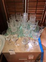 Group of clear crystal glasses displayed on a table, including various sizes and styles of water and wine glasses. Glasses are dusty.