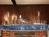 Full view of the assortment of glassware including candlesticks, wine glasses, dessert bowls, and other glass pieces on a blue cloth covered shelf against wood paneling