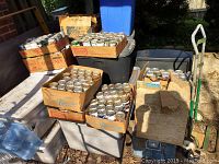 Multiple cardboard boxes and plastic bins filled with unopened vintage empty beer cans outdoors on a dolly and table.