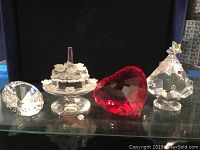 Clear crystal diamond shape, birthday cake with missing candle flame, red heart, and decorated Christmas tree figurines displayed on a glass shelf.