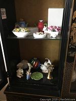 Overview of a display cabinet shelf featuring a collection of porcelain and brass decorative items including small floral pieces and teapots