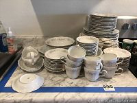 Full set of Johann Haviland china pieces including plates piled in stacks, cups arranged in front, and a creamer on a matching plate. White china with floral pattern and silver trim.