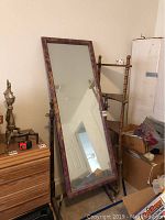 Side angled view of the antique stand-up mirror with purple and gold paint frame leaning against wall, next to wooden corner rack and drawers.