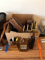 Box of vintage desk items seen from front including pen holders, clock, and brass items