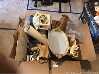 View of the box containing various home decor items including a white decorative birdcage, a brown woven purse, framed prints, rolled paper, and a small oval mirror tray.