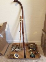 Wooden cane, eagle flagpole topper, Kiwanis bell, 86th Airborne Group brass pan, small figurine, and other small brass and metal items displayed in cardboard box and on floor.