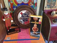 Photo of Beefeater Gin bottle holder with guard figure beside The Loser liquor dispenser and Pabst Blue Ribbon round wall sign on pink cloth.