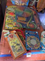 Seven vintage marble bagatelle games and Chinese Checkers game displayed together on bubble wrap surface. Some are shrink wrapped, colorful retro designs.