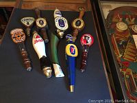 Photo showing nine vintage beer tap handles arranged on a dark surface. Includes brands Heineken, Whitbread, Newcastle Brown Ale, Bass, Fosters and McEwans.