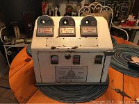 Front and top view of white metal American Postmaster stamp dispenser with three separate coin/stamp selector slots, showing paint chips and wear.