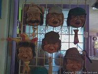 Six vintage spitter faces hanging on metal grid background in room with natural light. Faces have pull ties and visible water reservoirs in back. All faces show distinct expressions and hats.