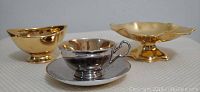 Front view of two gold lustre dishes and one silver lustre cup with saucer on neutral background