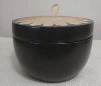 Front view of the round wooden storage box with black body and decorative lid showing the petal pattern and loop handle.