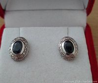 Pair of sterling silver stud earrings with black oval cabochon gemstones in beaded settings displayed on white cushion inside red box.