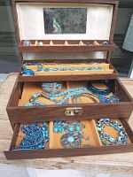 Jewelry box open showing three trays filled with blue and turquoise colored jewelry pieces including necklaces, bracelets, rings, and earrings arranged inside the box.