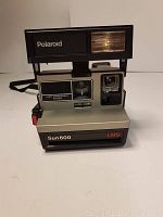 Front-facing view of the Polaroid Sun 600 LMS camera showing flash, lens, viewfinder, control buttons and strap.