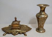 Front view of brass utility tray with ornate raised design and central square container topped with a pointed finial, and brass vase with etched floral pattern standing beside it.