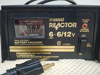 Close-up photo of the front panel of Unival Reactor 2 battery charger showing analog amperes meter, dual voltage indication (6/12V), and various labels including polarity check and overload protection.