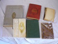 Seven vintage and antique books displayed on a flat surface, showing various sizes and covers including floral design, red, green, gray, beige, and one wrapped in plastic.