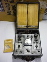 Top view of vintage tube tester with multiple tube sockets and analog meter alongside tube substitution handbook.