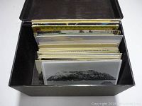 Photo showing black storage box filled with assorted vintage and antique postcards standing upright inside.